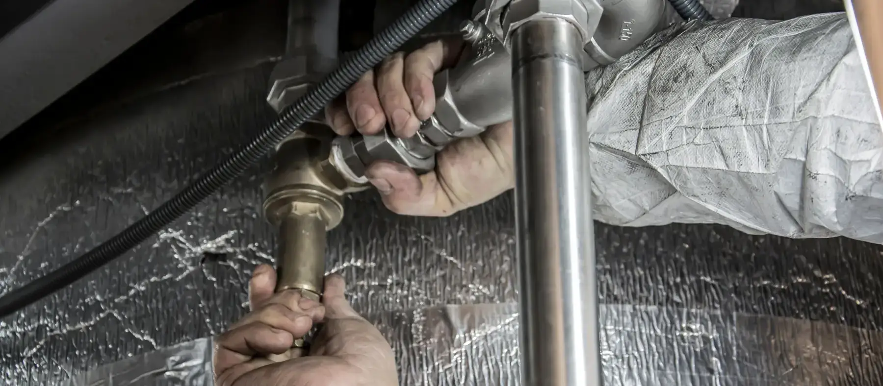 Hands tightening a threaded fitting on a steel pipe beside foil-wrapped insulation