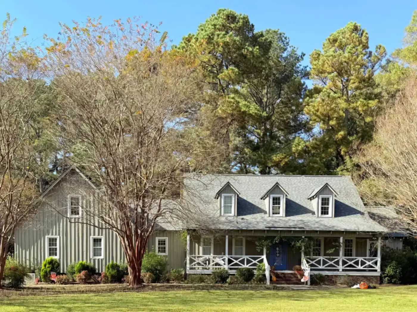 Gray country-style home with dormer windows and a white front porch among mature trees
