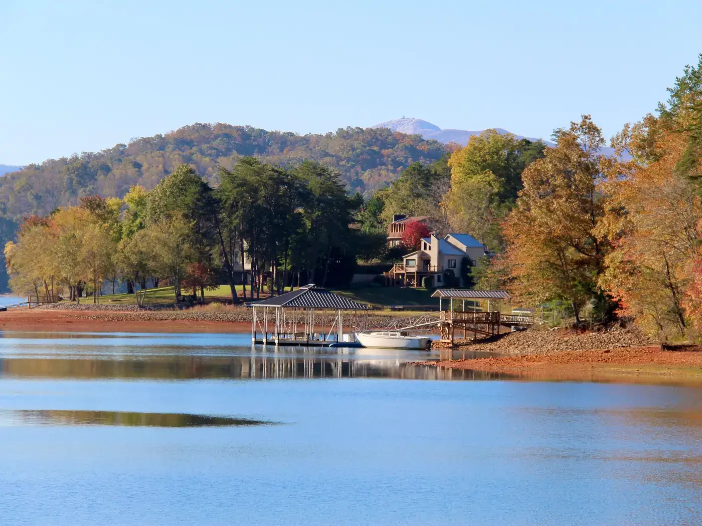 Lake homes and boat docks along a wooded shoreline with autumn trees and mountains beyond