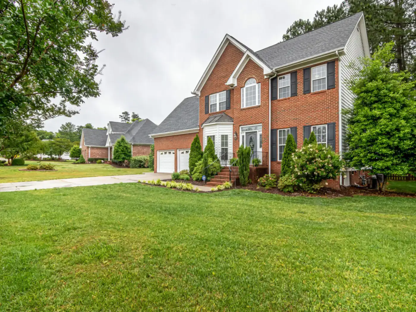 Two-story brick colonial home with black shutters on a large lawn in a tree-lined neighborhood